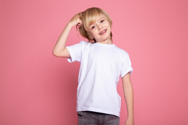 Blonde smiling boy in white t-shirt and grey jeans on pink wall