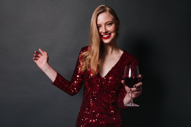 Blonde smiling woman drinking red wine. Studio shot of pretty girl in dress dancing on black background.