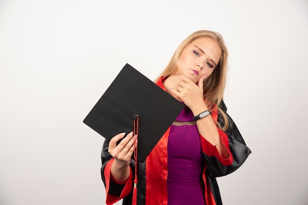 Blonde student holding her cap while thinking on white wall.