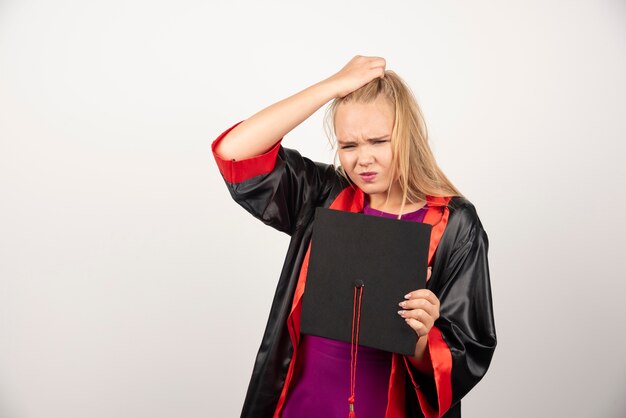 Blonde student holding her cap while thinking on white wall. 