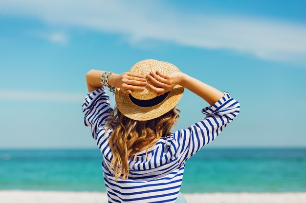 Блондинка стильная женщина в соломенной шляпе и солнцезащитных очках, позирует на райском тропическом пляже