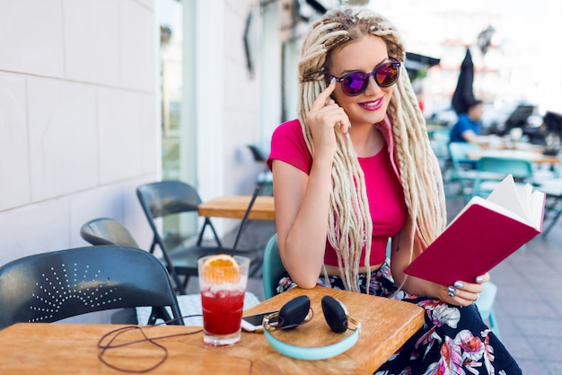 Blonde unusual woman with dreadlocks sitting in cafe on the street, holding notebook, enjoying free time. Wearing bright pants with tropical print.