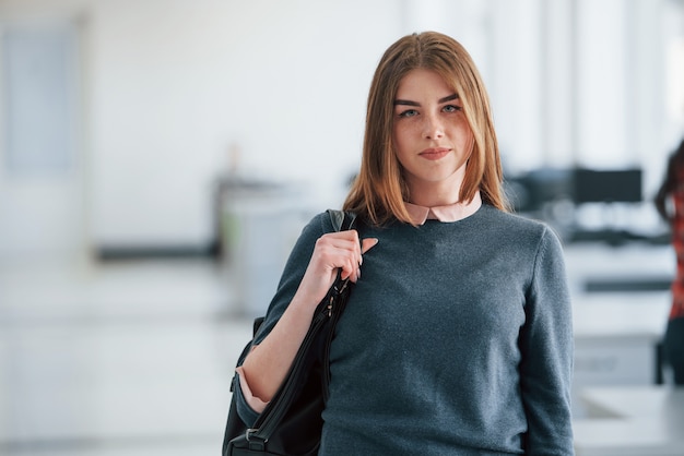 Blonde with black bag in hands. Portrait of attractive young woman standing in the office