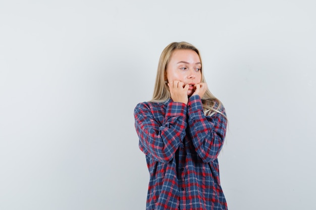 Blonde woman biting fingers in checked shirt and looking menaced , front view.
