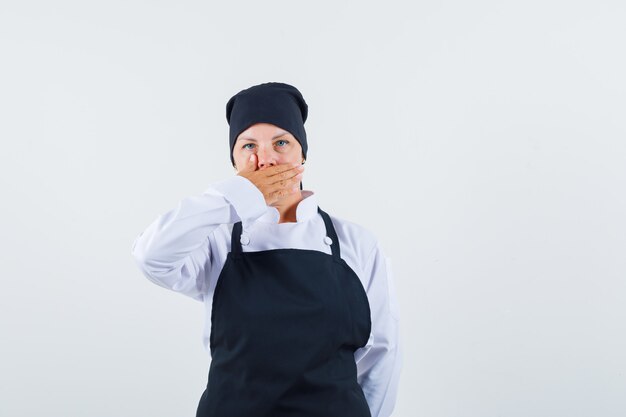 手で口を覆い、驚いて見える、正面図の黒い料理の制服を着た金髪の女性。