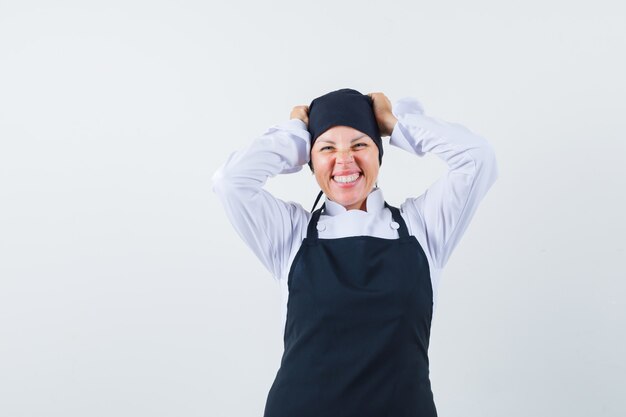 Blonde woman in black cook uniform holding hands on head, grimacing and smiling gracefully and looking pretty