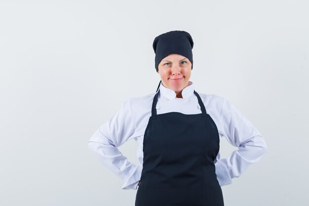 Blonde woman in black cook uniform holding hands behind waist and looking pretty