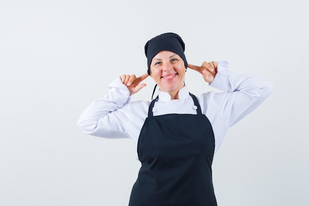 Blonde woman in black cook uniform plugging ears with index fingers and looking pretty , front view.