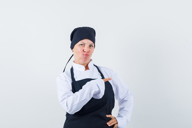 Blonde woman in black cook uniform pointing right with index finger,curving lips and looking pretty