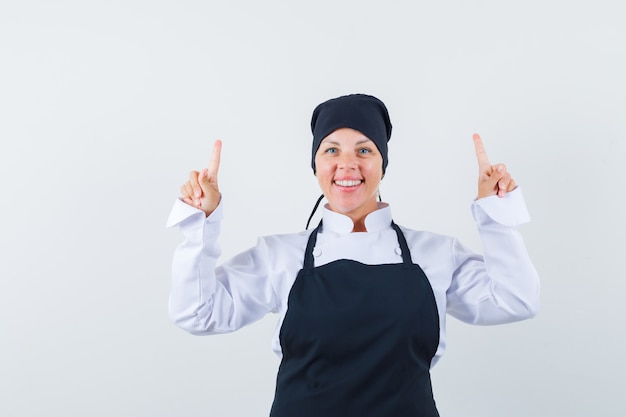 Blonde woman in black cook uniform pointing up with index fingers and looking pretty