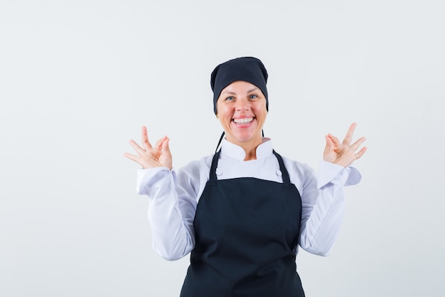 Donna bionda in uniforme nera del cuoco che mostra i segni giusti con entrambe le mani e che sembra felice, vista frontale.