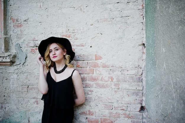 Free photo blonde woman on black dress necklaces and hat against old wall at street of city