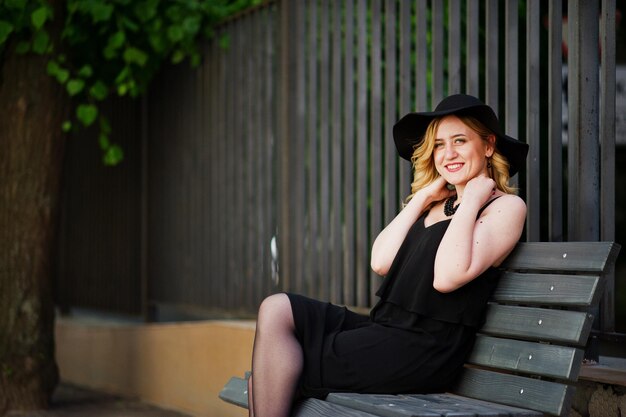 Blonde woman on black dress necklaces and hat sitting on bench