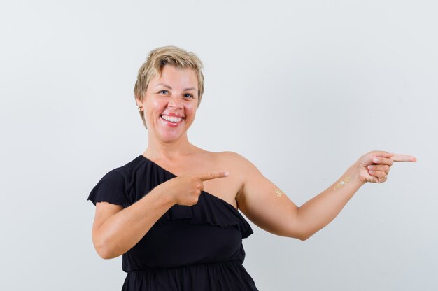 Blonde woman in black dress pointing right with index fingers and smiling and looking optimistic