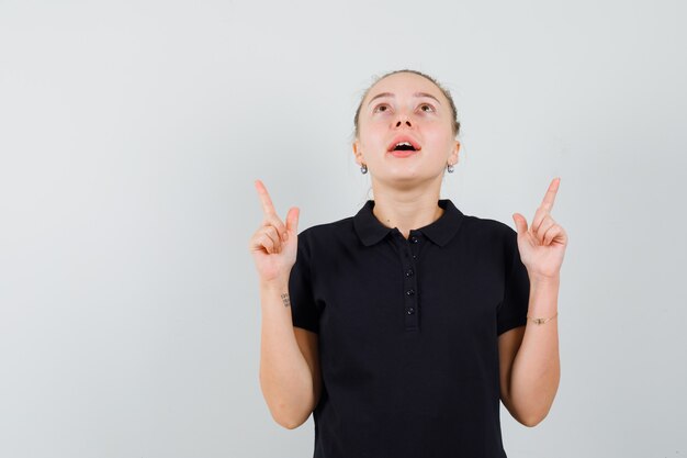 Blonde woman in black t-shirt pointing upwards and looking upward and looking optimistic