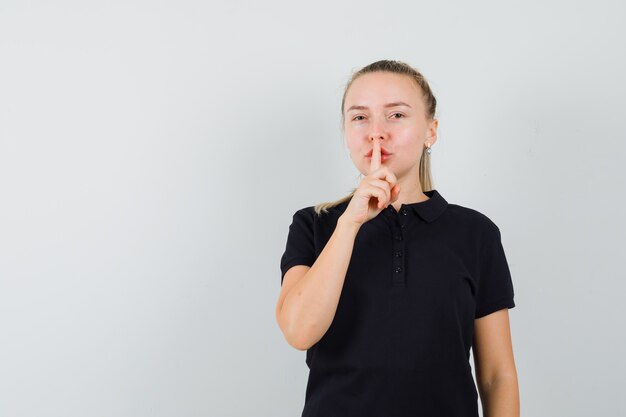 Blonde woman in black t-shirt showing silence gesture and looking happy