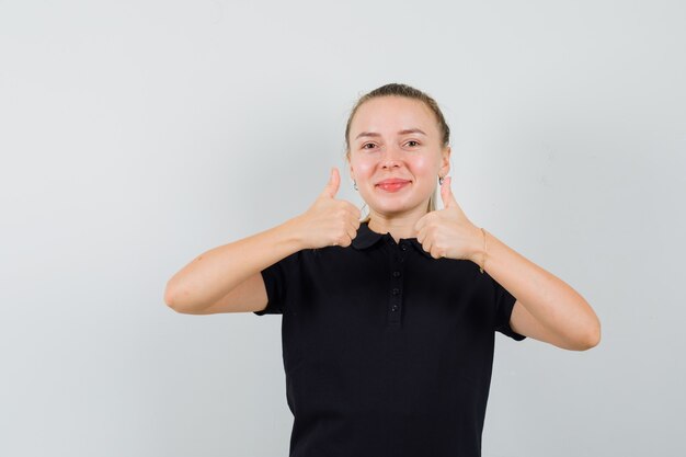 彼女の両手で親指を示し、笑顔で幸せそうに見える黒いTシャツのブロンドの女性