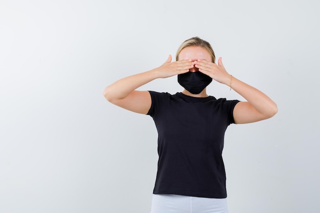 Foto gratuita donna bionda in maglietta nera, pantaloni bianchi, maschera nera che copre gli occhi con le mani