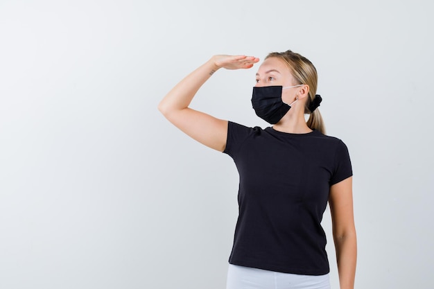 Blonde woman in black t-shirt, white pants, black mask looking far away