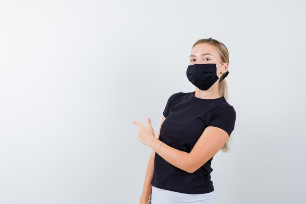 Blonde woman in black t-shirt, white pants, black mask pointing left