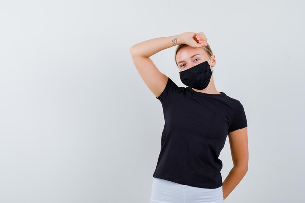 Blonde woman in black t-shirt, white pants, black mask raising arm over head