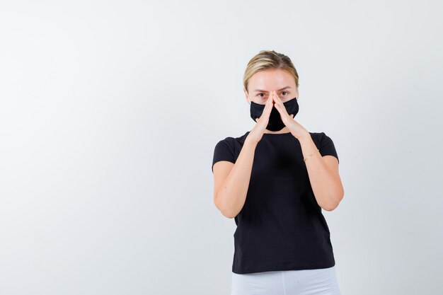 Blonde woman in black t-shirt, white pants, black mask showing insurance gesture