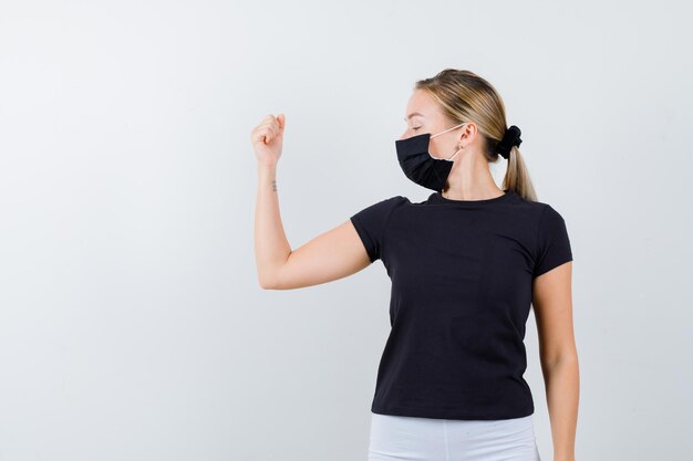 Blonde woman in black t-shirt, white pants, black mask showing muscles