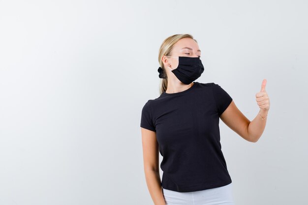 Blonde woman in black t-shirt, white pants, black mask showing thumb up