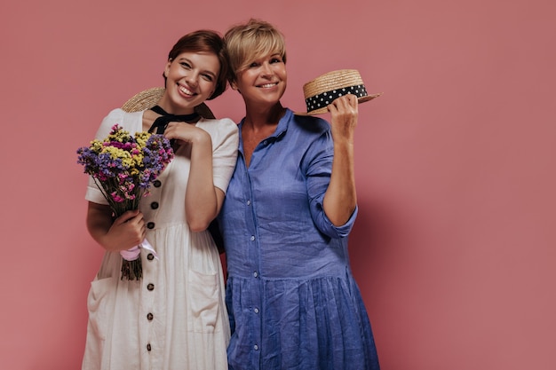 Foto gratuita donna bionda in vestito blu che tiene il cappello di paglia e sorridente con una ragazza dai capelli corti in abiti leggeri con fiori di campo colorati su sfondo rosa.