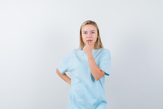 Blonde woman in blue t-shirt holding one hand behind waist, putting index finger on chin