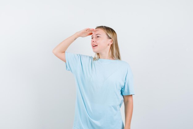 Blonde woman in blue t-shirt looking far away with hand over head and looking focused