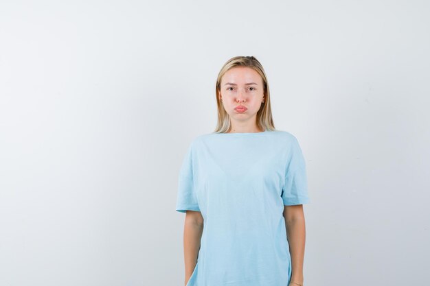 Blonde woman in blue t-shirt puffing cheeks while posing and looking disappointed