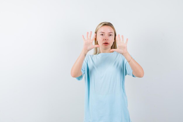 Blonde woman in blue t-shirt raising palms in surrender gesture and looking cute