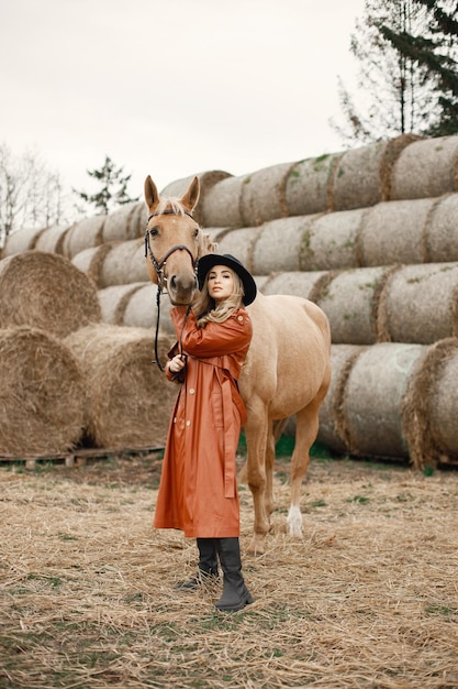 Blonde woman and brown horse standing on a farm near hay bales. Woman wearing black dress, red leather coat and hat. Woman touching the horse.