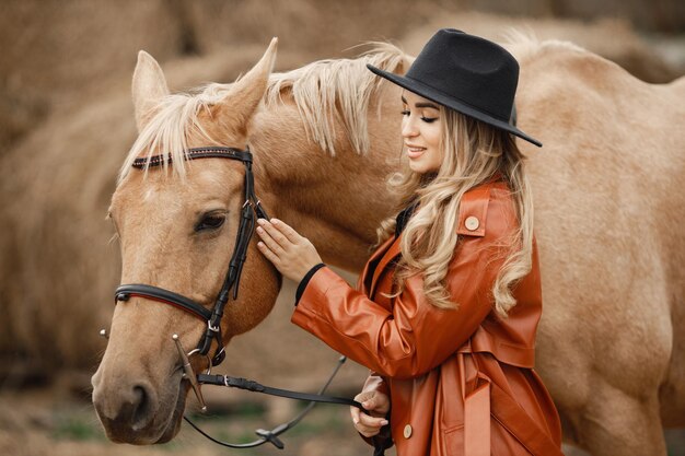Blonde woman and brown horse standing on a farm near hay bales. Woman wearing black dress, red leather coat and hat. Woman touching the horse.