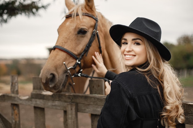 Blonde woman and brown horse standing on a farm. Woman wearing black clothes and hat. Woman touching the horse behinde the fence.
