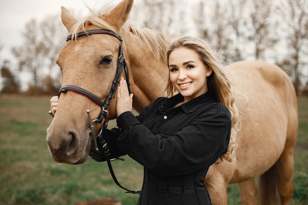 Блондинка и коричневая лошадь стоят в поле. Женщина в черной одежде. Женщина трогает лошадь.