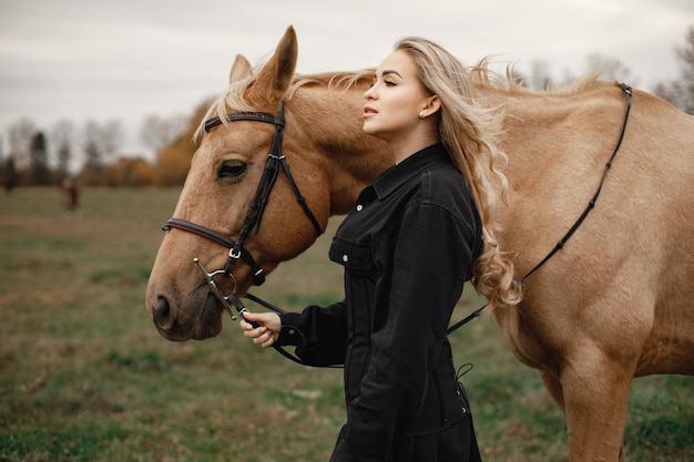 Foto gratuita donna bionda e cavallo marrone in piedi nel campo. donna che indossa abiti neri. donna che tocca il cavallo.