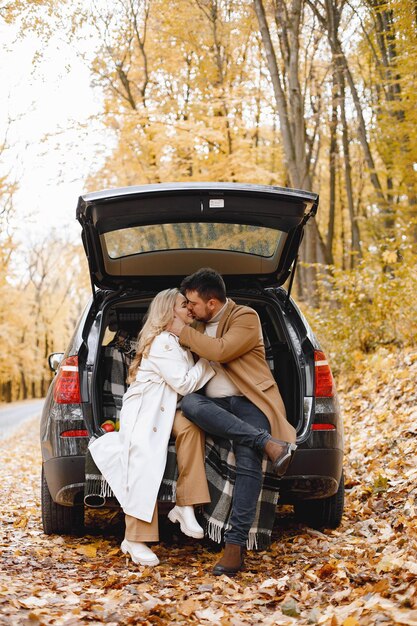 금발 여자와 갈색 머리 남자가 숲에서 검은 차 트렁크에 앉아. 흰색 코트와 남자 베이지색 코트를 입고 여자입니다. 사랑스러운 커플 키스.