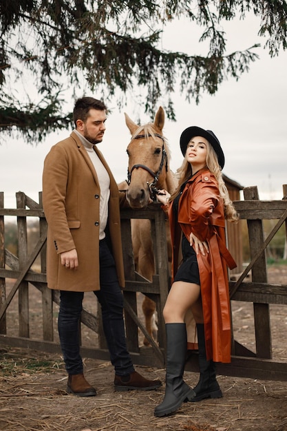 Foto gratuita donna bionda, uomo bruna in piedi in una fattoria vicino al recinto con cavallo marrone. donna che indossa abiti neri e cappotto di pelle rossa e uomo che indossa un cappotto marrone. uomo e donna che toccano il cavallo