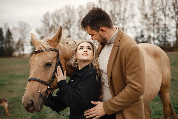 Blonde woman, brunette man standing in the field with brown horse. Woman wearing black clothes and man wearing brown coat. Man and woman hugging.