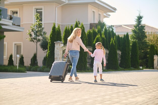 Blonde woman carrying a suitcase and holding her daughters hand