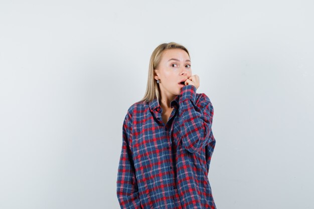 Blonde woman in checked shirt biting fingers and looking scared , front view.