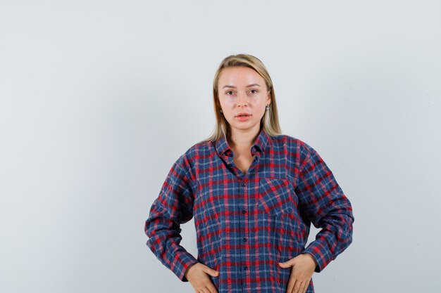 Blonde woman in checked shirt holding hands on belly, posing at camera and looking charming , front view.