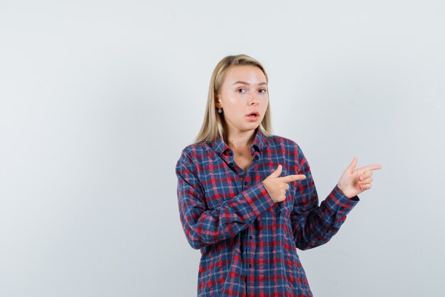 Blonde woman in checked shirt pointing right with index fingers and looking surprised , front view.