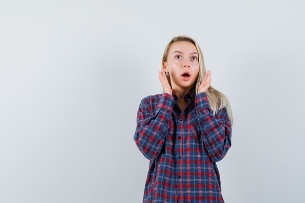 Blonde woman in checked shirt putting hands near mouth and looking surprised , front view.