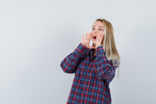 Blonde woman in checked shirt putting hands near mouth and shouting and looking attractive , front view.