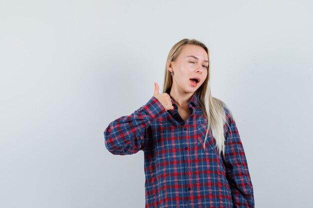 Blonde woman in checked shirt showing thumb up and winking and looking attractive , front view.