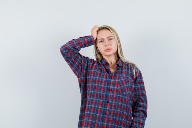 Blonde woman in checked shirt standing in thinking pose, holding hand on head and looking pensive , front view.