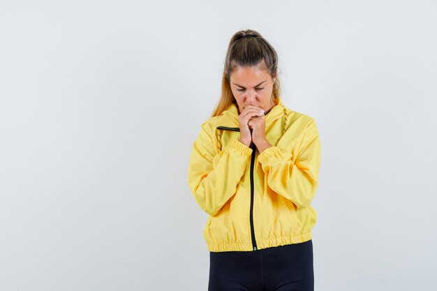 Blonde woman clenching fists and standing in prayer pose in yellow bomber jacket and black pants and looking focused